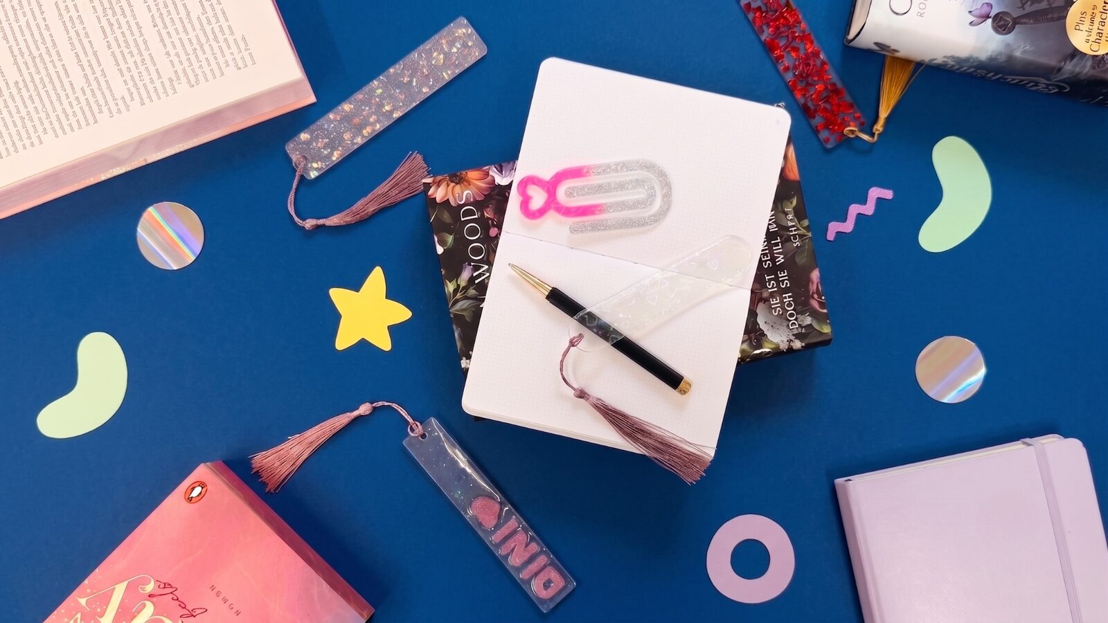 Selbstgemachte Lesezeichen aus UV-Resin mit Glitzer, Quasten und eingegossenen Motiven, arrangiert mit Büchern und Notizbuch
